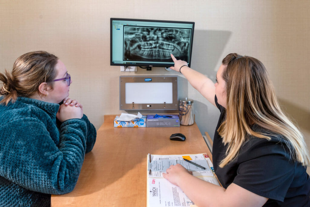 A dental professional explaining a patient's X-ray displayed on a monitor, while the patient listens attentively, seated at a desk with forms and documents spread out in front of them.