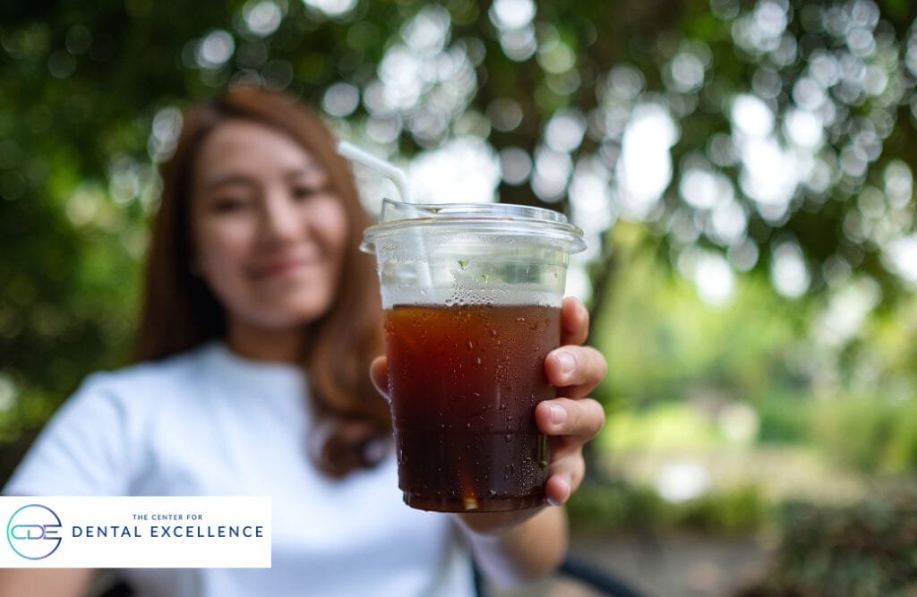 Smiling woman holding a cup of iced coffee outdoors, illustrating beverages that can cause dental staining and affect oral health.